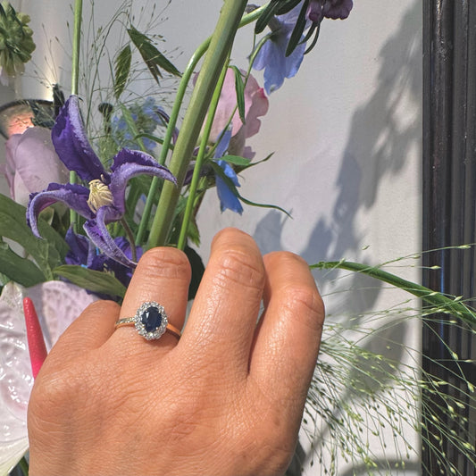 An oval sapphire and round diamond cluster ring in yellow and white gold, modelled on a ring finger, with flowers in the background