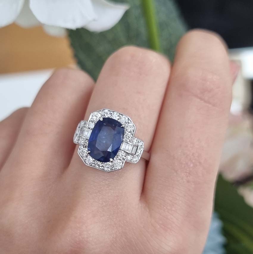 3.3ct Sapphire and Diamond Cluster Dress Ring, 3.30ct oval blue sapphire surrounded by round brilliant-cut diamonds with brilliant and baguette-cut diamond buckle style shoulders in 18ct white gold