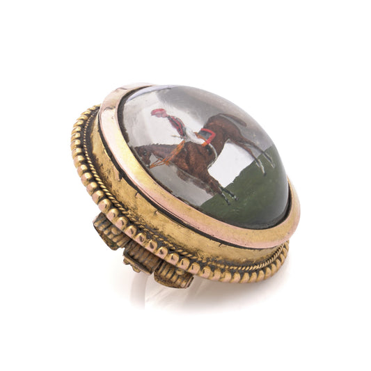 A round crystal brooch in yellow gold featuring a jockey sitting on a horse's back, on a white background