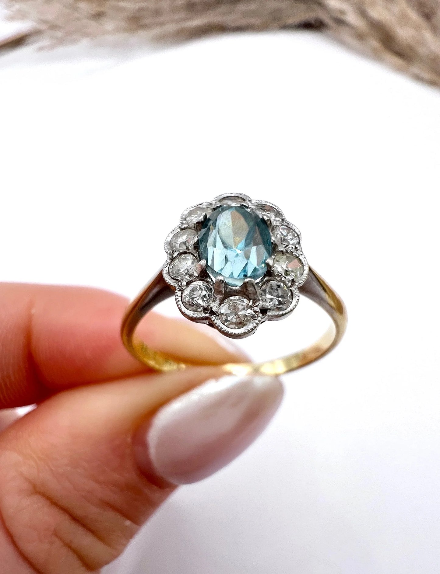 An oval cluster ring with a blue zircon centre surrounded by round diamonds, in yellow gold and platinum, held in a hand