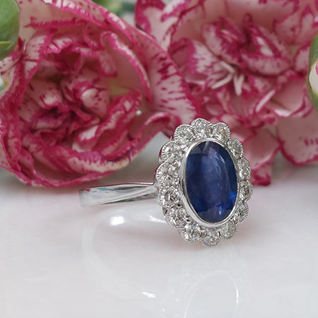 An oval sapphire and brilliant cut diamond cluster ring in white gold, with pink flowers in the background