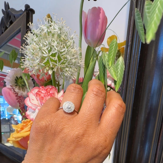 A diamond cluster ring in white gold, modelled on a hand, with flowers in the background