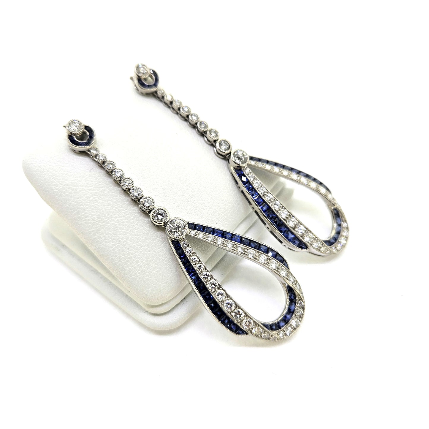 A pair of sapphire and diamond teardrop shaped long drop earrings in white gold, on an earring display pad, against a white background