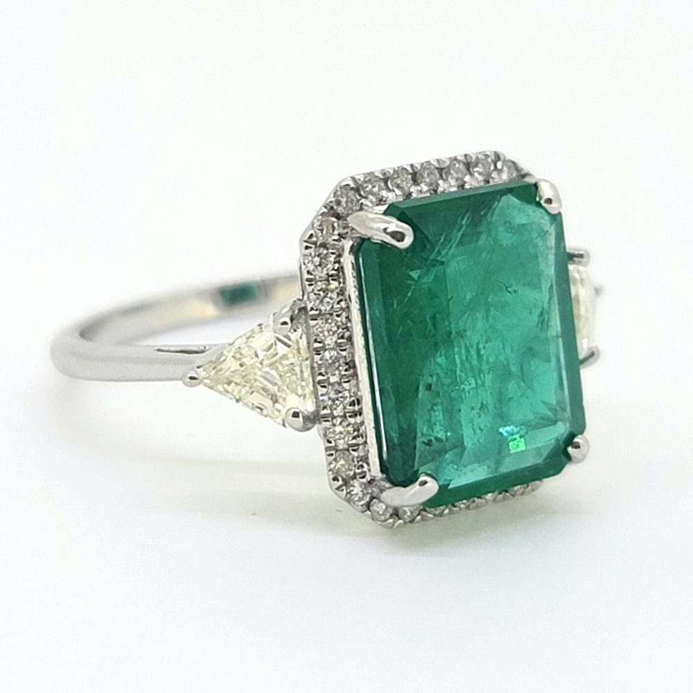 An emerald and diamond octagonal shaped cluster ring with trilliant cut diamond shoulders in white metal, against a white background