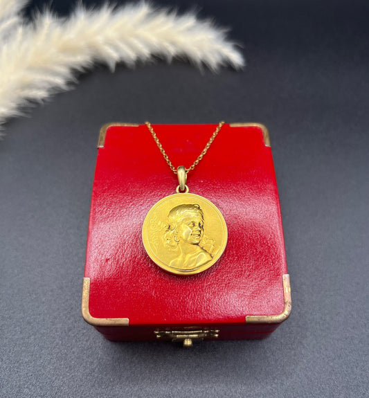 A round gold pendant featuring a girl with the text AMOR, on a chain, on a red antique box