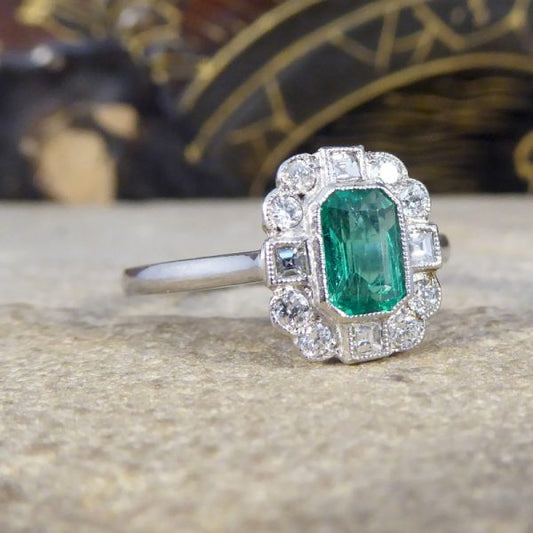 A beautiful contemporary ring featuring a 0.50ct emerald cut emerald with a diamond surround. With princess and modern brilliant cut diamonds alternating around the emerald, this ring has a total of 0.35ct diamonds allowing it to sparkle beautifully on the finger all set in platinum in a millegrain edge.