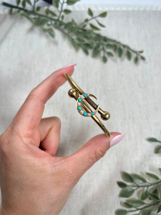 A yellow gold crossover bangle bracelet with a pearl and turquoise crescent moon centre, held in a hand