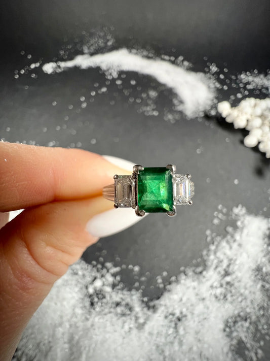 A three stone emerald and diamond ring in platinum, held in a hand