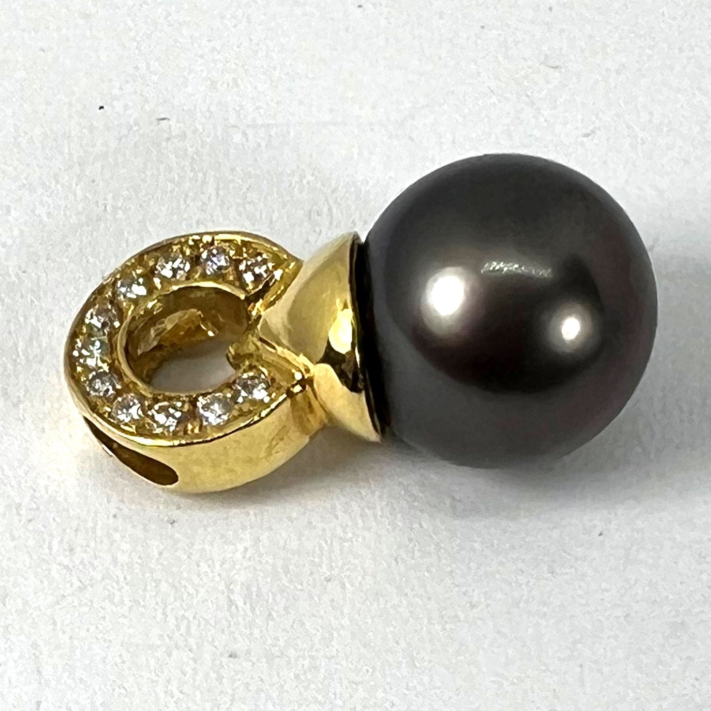 A black pearl pendant with a yellow gold bale set with diamonds, on a white background