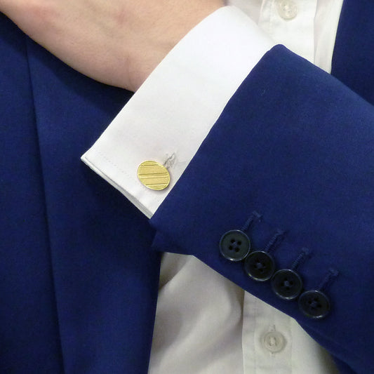 A yellow gold oval cufflink with engraving on a white shirt cuff, against a royal blue suit