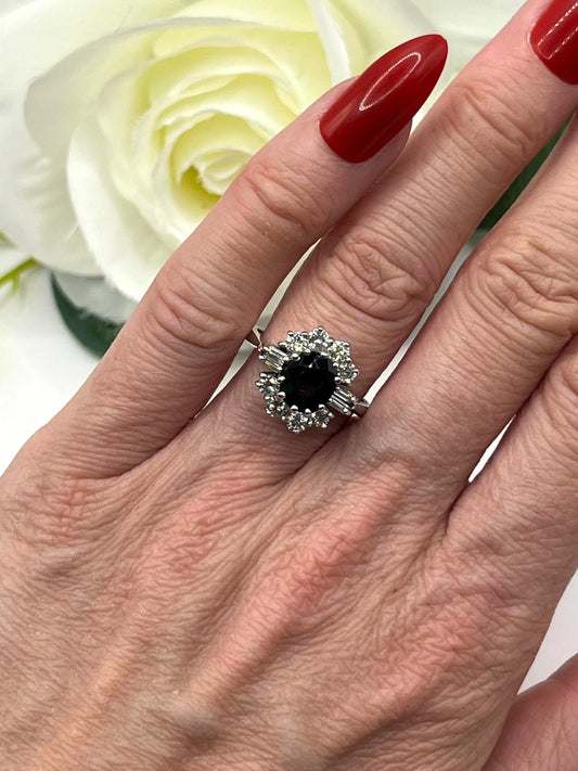 An oval cluster ring with a sapphire centre, round diamond surround and baguette cut diamond shoulders in white gold, modelled on a hand