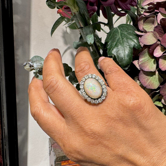 An oval shaped cluster ring with a central opal surrounded by diamonds, in gold and silver, modelled on a hand