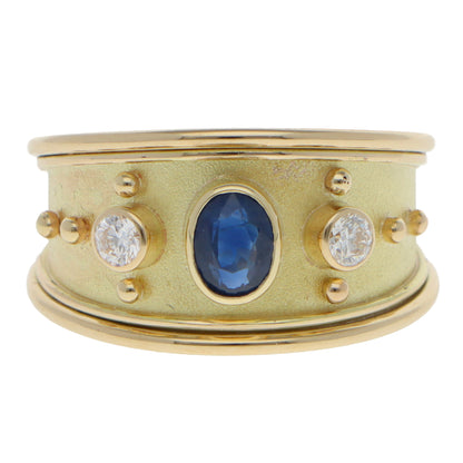 A yellow gold three stone band ring with an oval blue sapphire and two round diamonds, on a white background