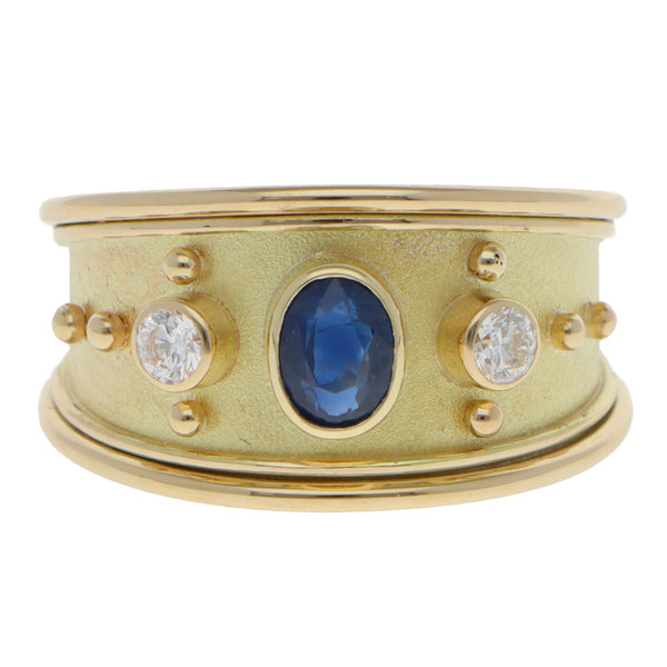A yellow gold three stone band ring with an oval blue sapphire and two round diamonds, on a white background