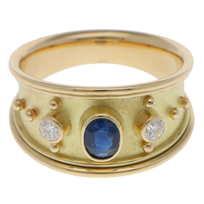 A yellow gold three stone band ring with an oval blue sapphire and two round diamonds, on a white background
