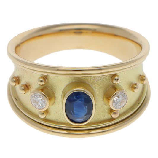 A yellow gold three stone band ring with an oval blue sapphire and two round diamonds, on a white background