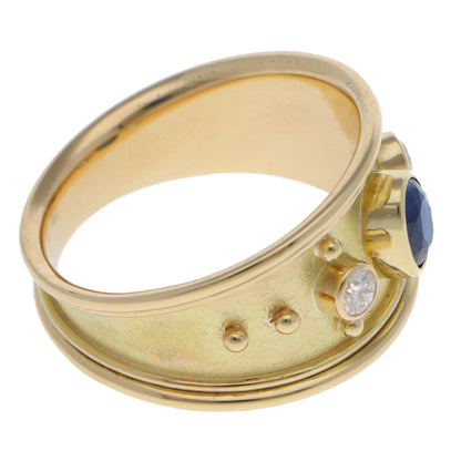 A yellow gold three stone band ring with an oval blue sapphire and two round diamonds, on a white background