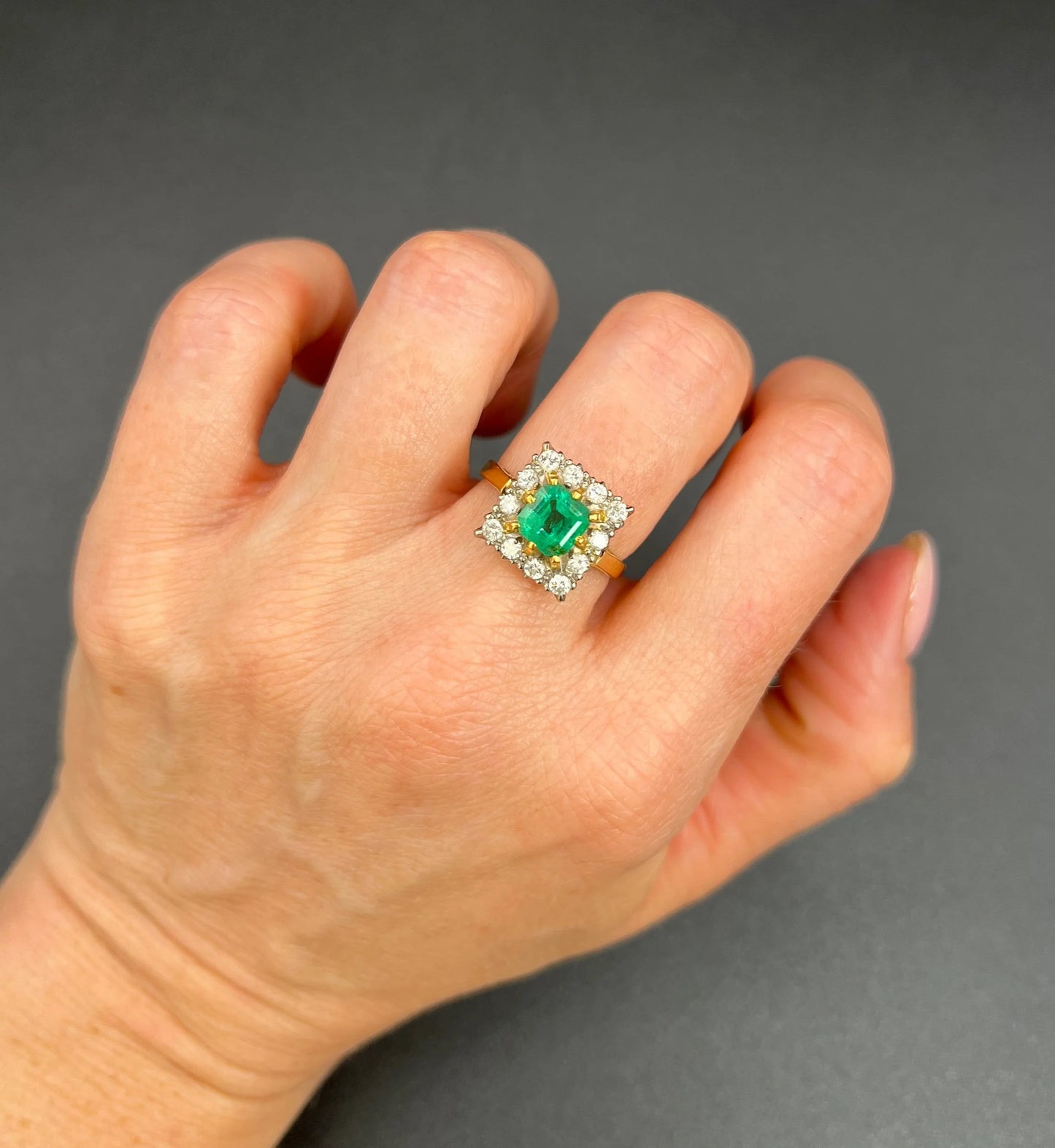 A square shaped emerald and diamond cluster ring in yellow and white gold, modelled on a hand