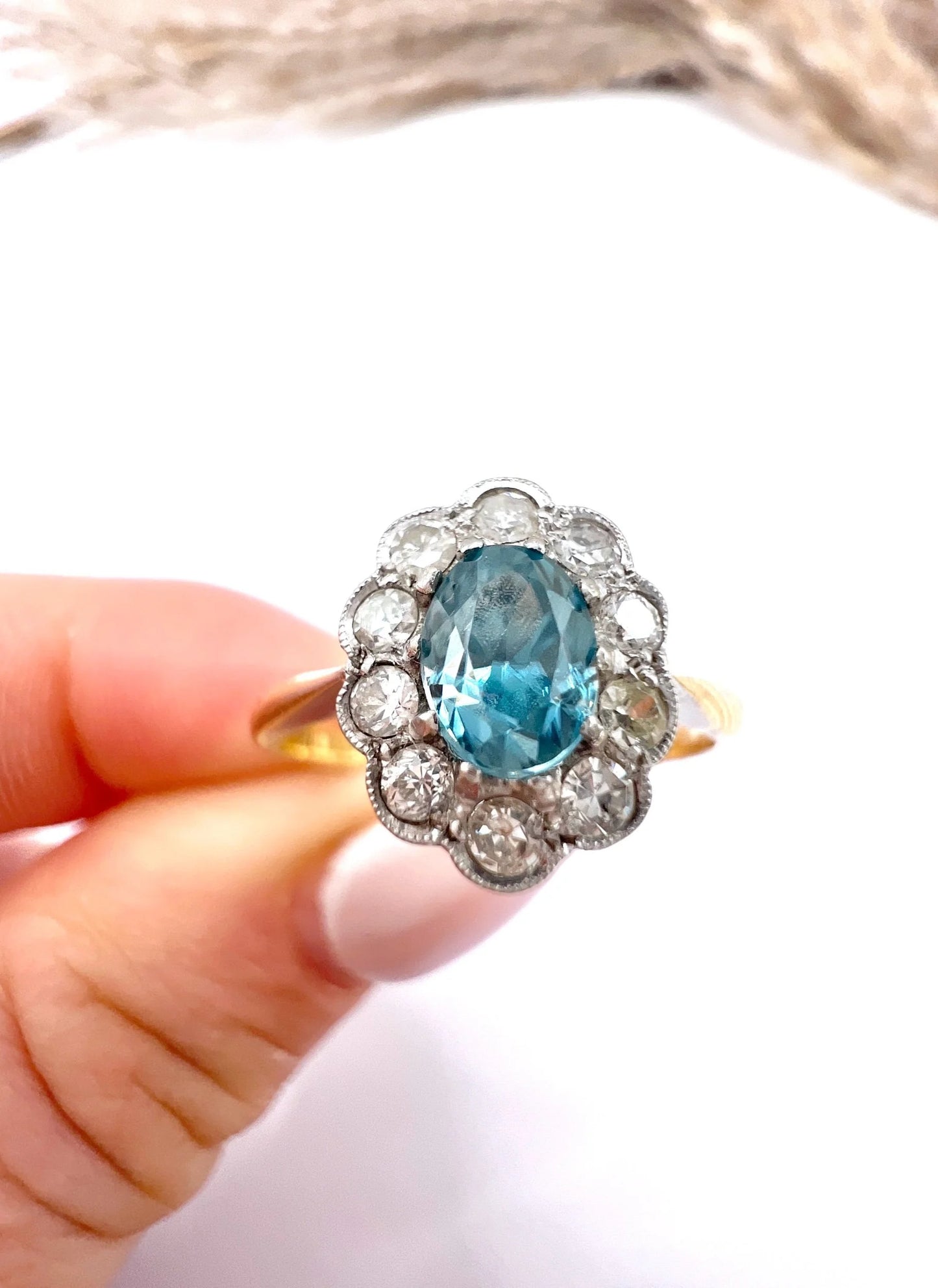 An oval cluster ring with a blue zircon centre surrounded by round diamonds, in yellow gold and platinum, held in a hand