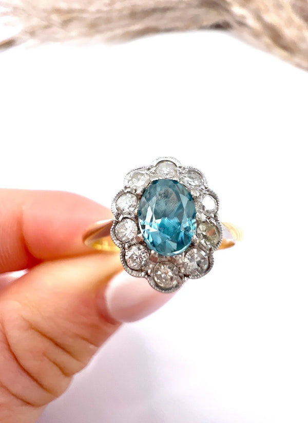 An oval cluster ring with a blue zircon centre surrounded by round diamonds, in yellow gold and platinum, held in a hand
