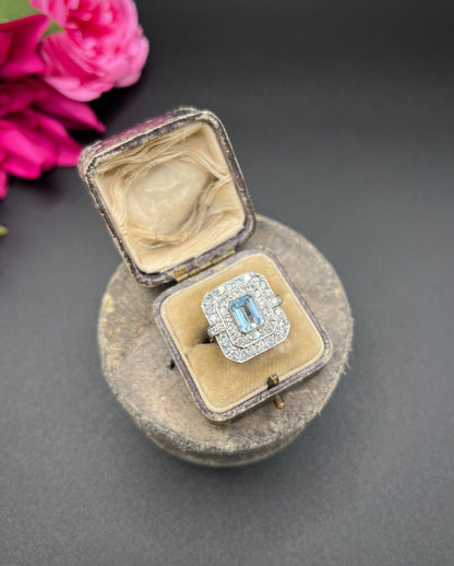An octagonal shaped cluster ring with an aquamarine centre surrounded by two rows of diamonds, in platinum, in a ring box