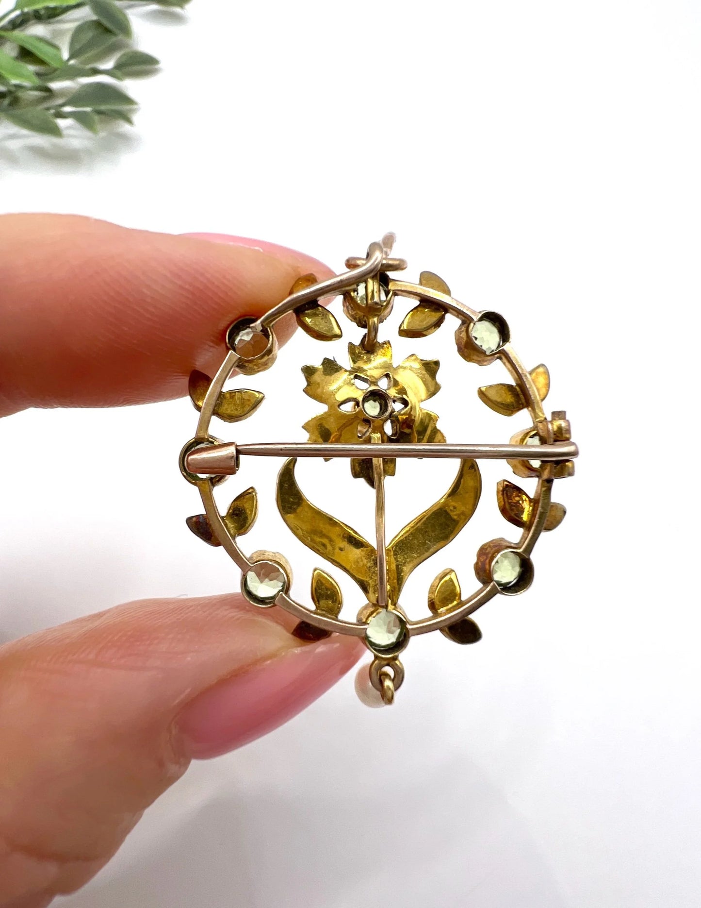 A peridot and pearl floral circle brooch/pendant in yellow gold, held in a hand