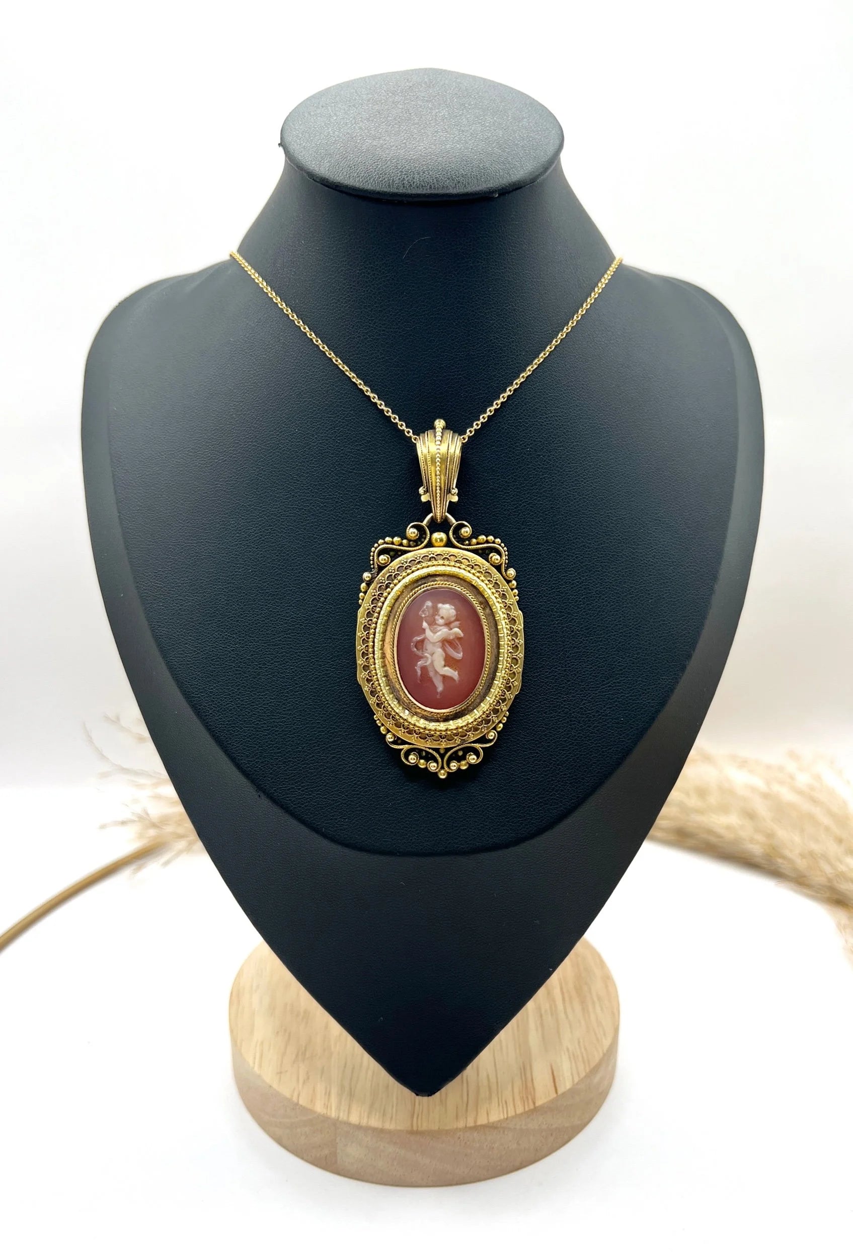 An ornate yellow gold oval cherub cameo locket pendant, with a chain, on a black jewellery bust