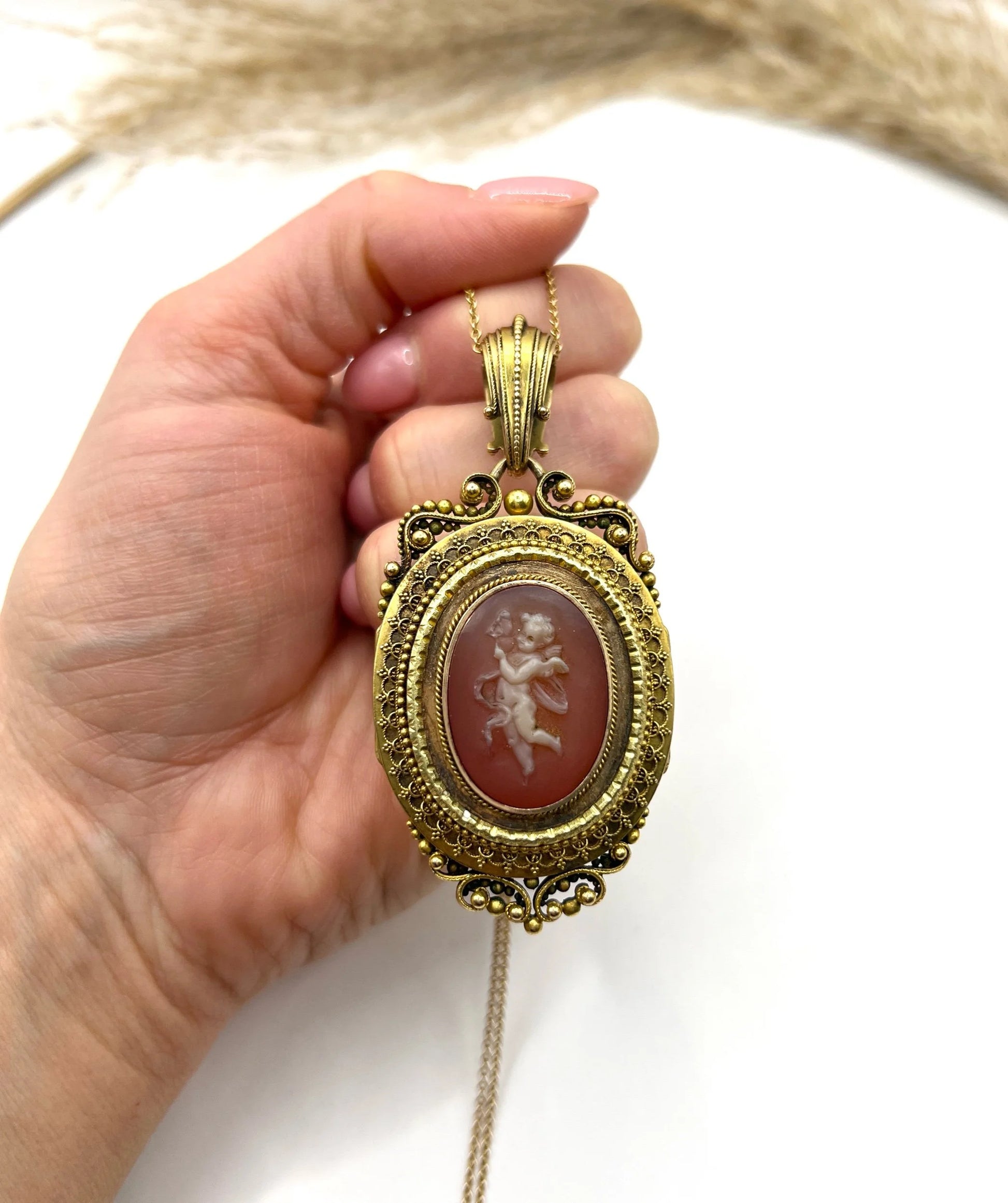 An ornate yellow gold oval cherub cameo locket pendant, with a gold chain, held in a hand