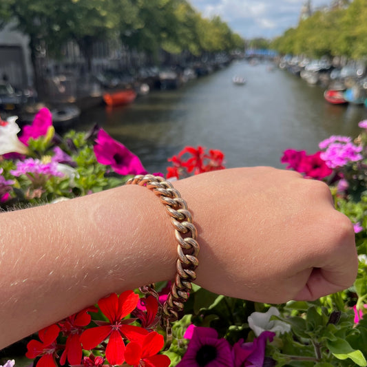 A yellow gold curb link bracelet with heart padlock clasp, modelled on a wrist against flowers and a canal