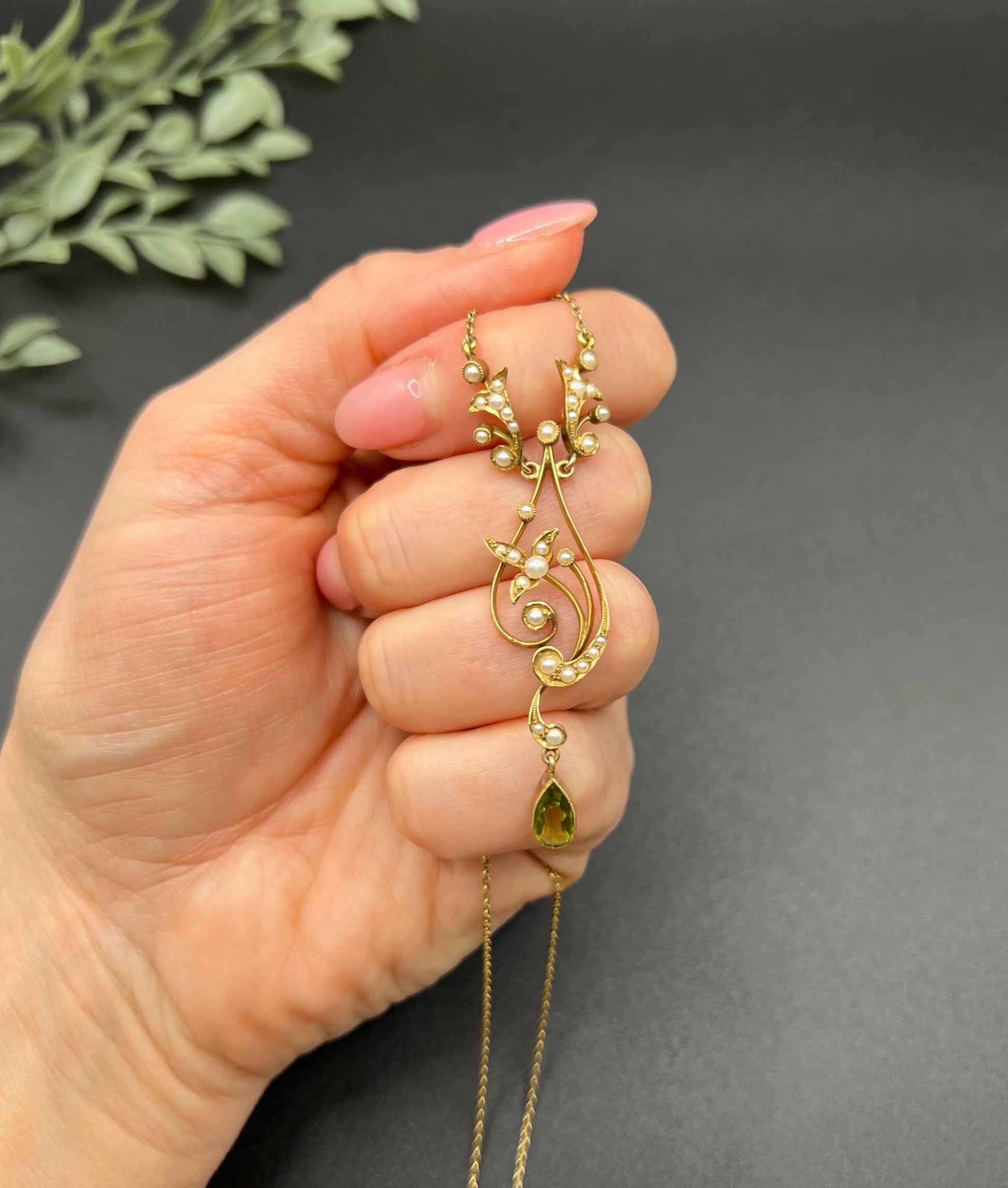 A yellow gold floral pendant necklace with pearls and a peridot drop, held in a hand
