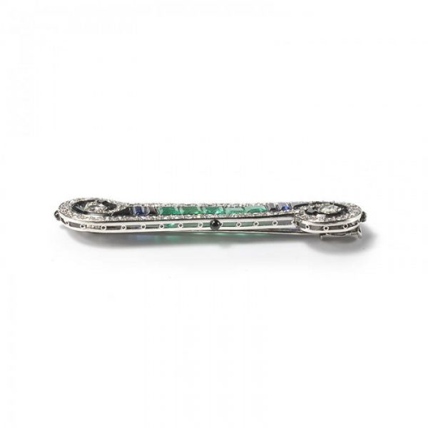 Art Deco French Emerald Sapphire Onyx and Diamond Bar Brooch in Platinum; set with 2.00cts calibre-cut emeralds, 0.50cts calibre-cut sapphires and fancy-cut black onyx, framed by 2.50cts old-cut and single-cut diamonds, in a scroll design, with enamelled detail