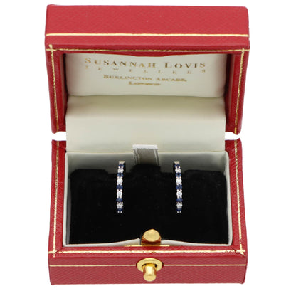 A pair of white gold hoop earrings set with round sapphires and diamonds, in a red earring box, on a white background