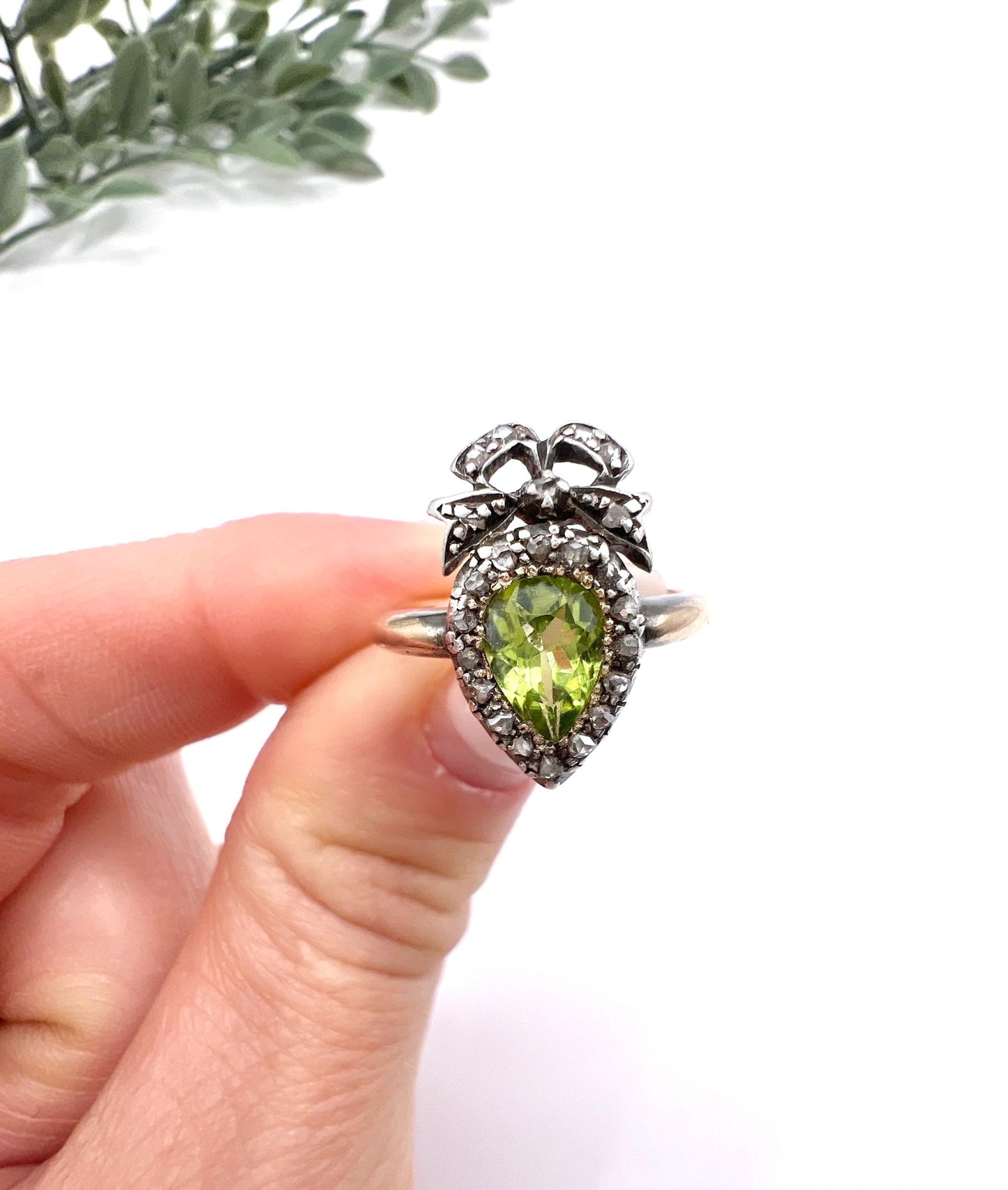 A pear cut peridot and round diamond cluster ring with bow detail, in yellow gold and white metal, held in a hand