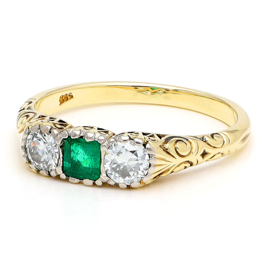 A yellow gold ring with a square cut emerald and two old cut diamonds, with scroll detailing, against a white background