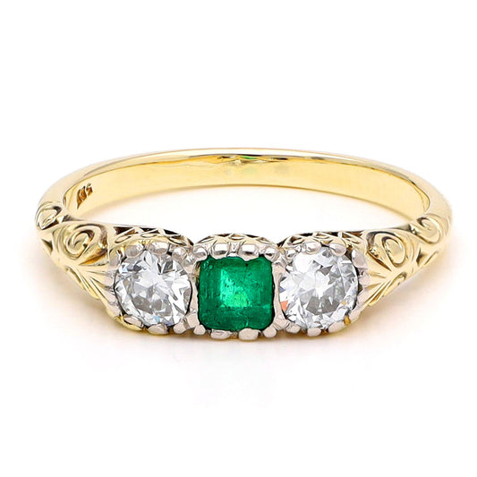A yellow gold ring with a square cut emerald and two old cut diamonds, with scroll detailing, against a white background