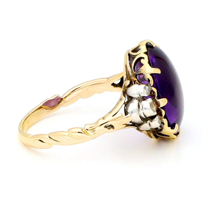 A buff top, oval shaped amethyst ring with rose cut diamond shoulders in yellow and white gold, against a white background