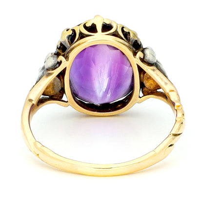 A buff top, oval shaped amethyst ring with rose cut diamond shoulders in yellow and white gold, against a white background