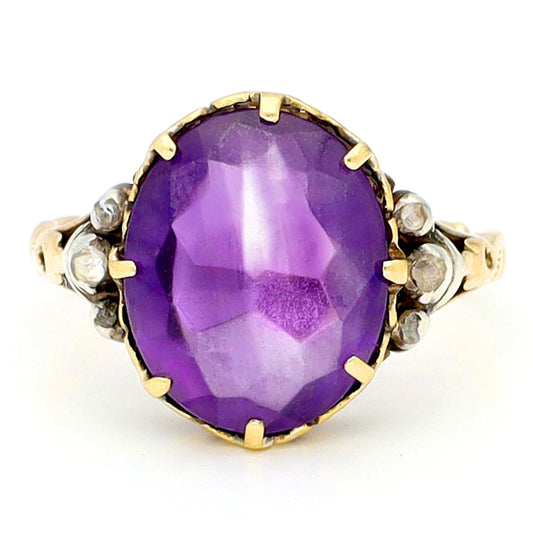 A buff top, oval shaped amethyst ring with rose cut diamond shoulders in yellow and white gold, against a white background