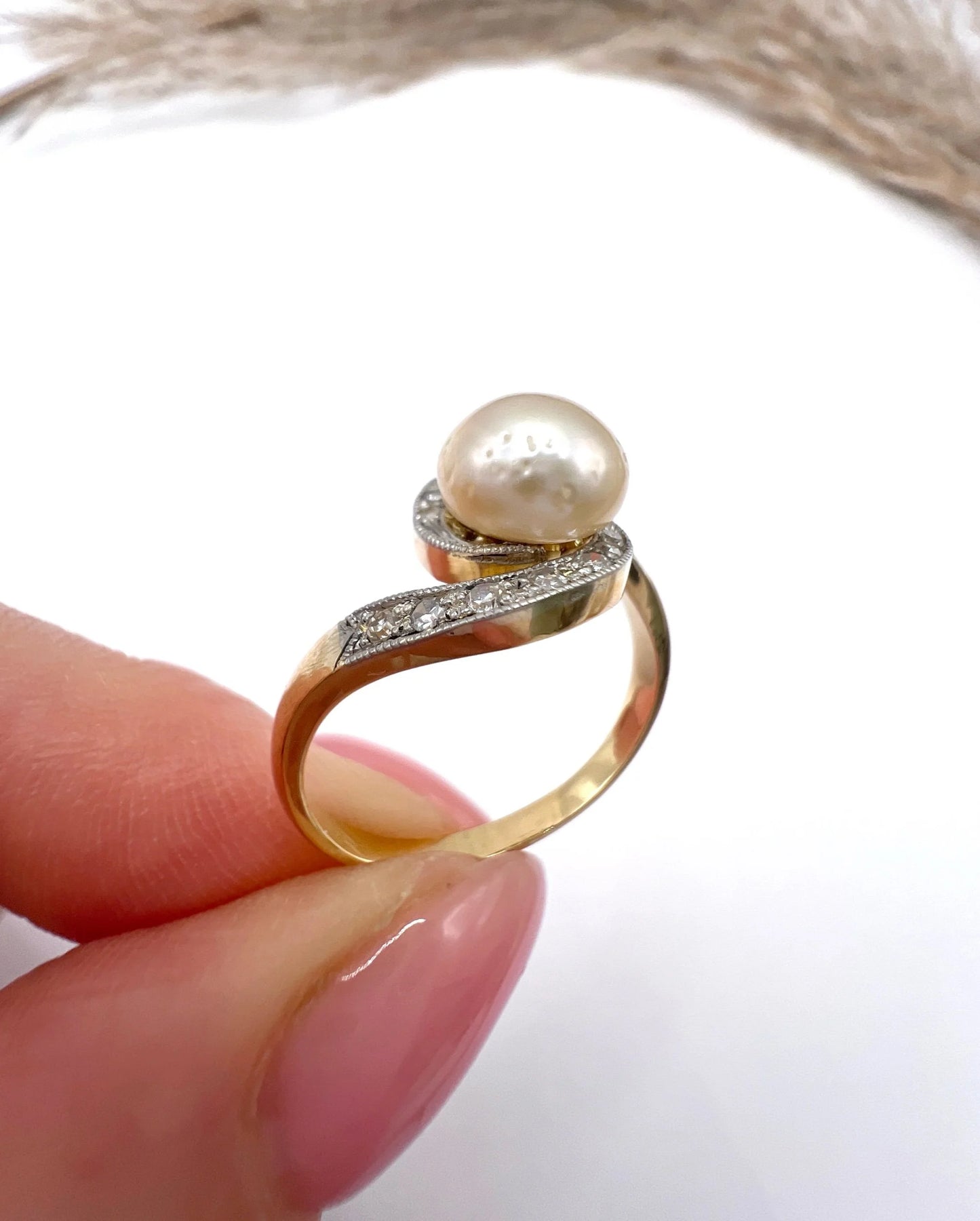 A pearl and diamond crossover swirl ring in yellow and white gold, held in a hand