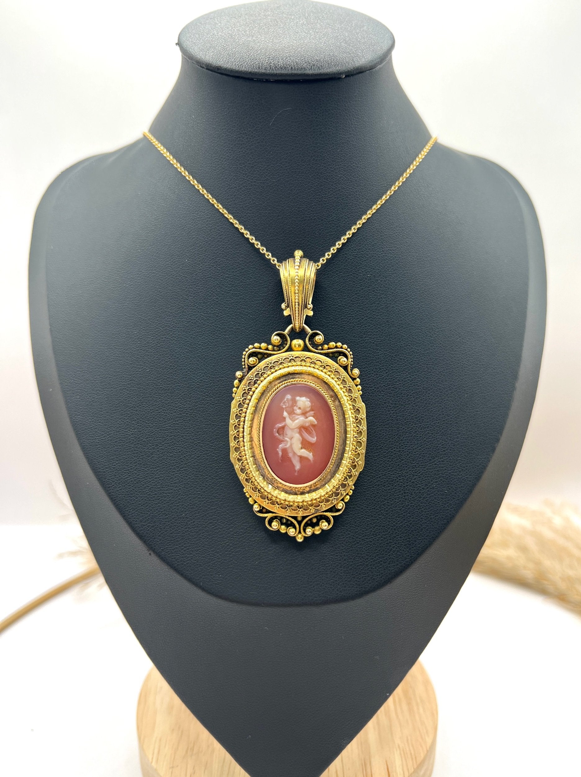 An ornate yellow gold oval cherub cameo locket pendant, with a chain, on a black jewellery bust