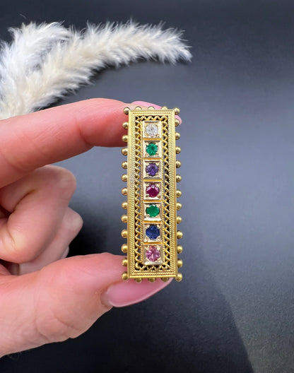A yellow gold multi-gemstone and diamond acrostic brooch, held in a hand