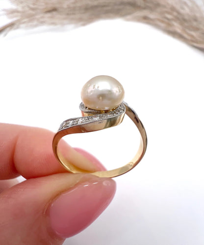 A pearl and diamond crossover swirl ring in yellow and white gold, held in a hand