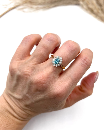 An oval cluster ring with a blue zircon centre surrounded by round diamonds, in yellow gold and platinum, modelled on a hand