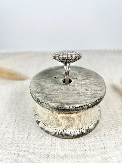 A navette shaped diamond cluster ring in white metal, sitting on top of a wooden and cloth object