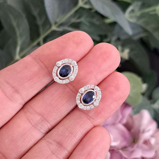 A pair of oval sapphire and round diamond cluster earrings in yellow and white gold, held in a hand