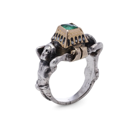 A silver and gold rennaisance ring styled as two men holding the setting, with a single square emerald, on a white background