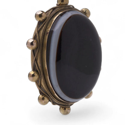 An oval pendant with a cabochon cut banded black agate with yellow gold hobnail bead border, on a white background
