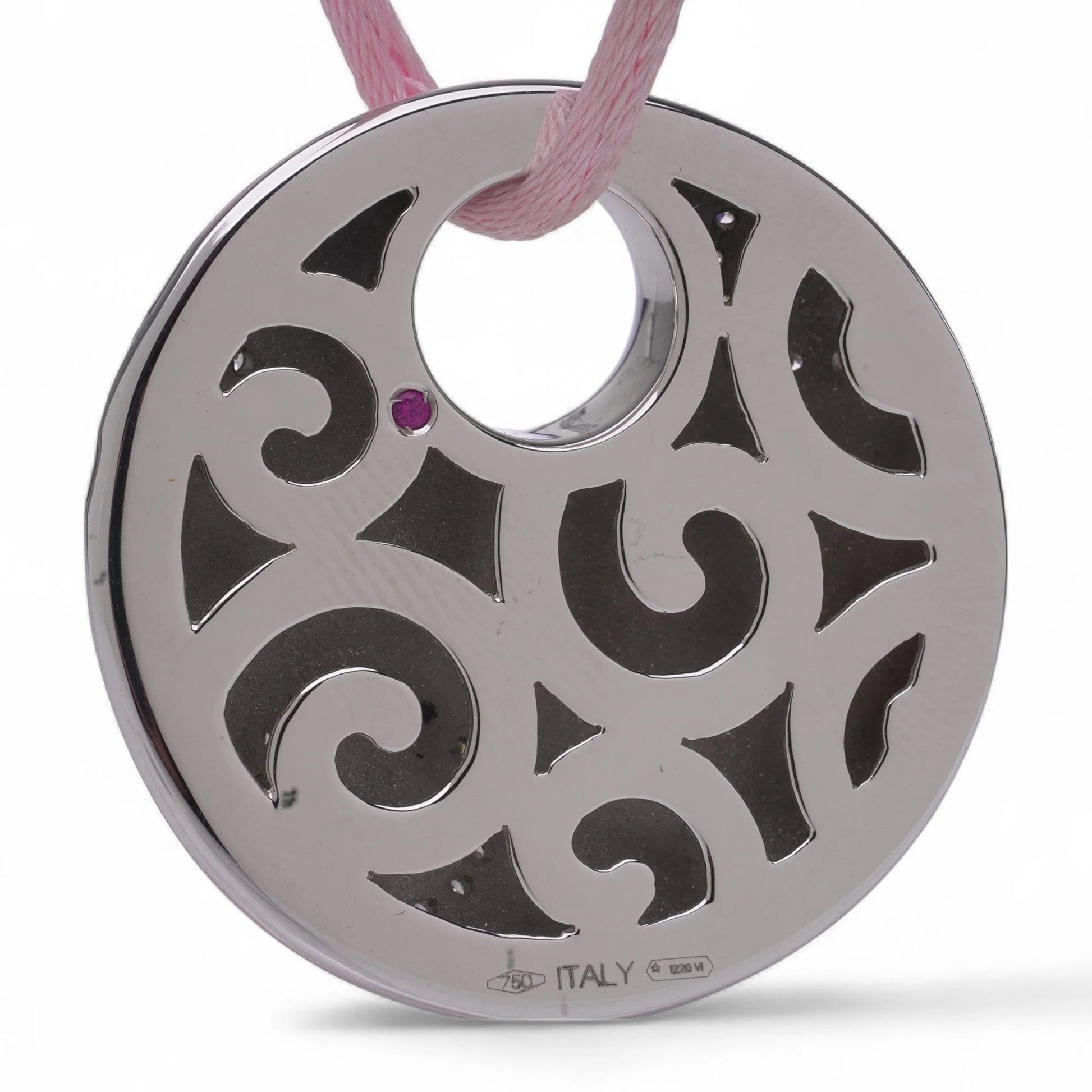 A white gold circular pendant with a cutout design and a pink sapphire, on a pink cord, against a white background
