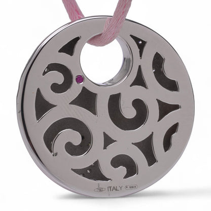 A white gold circular pendant with a cutout design and a pink sapphire, on a pink cord, against a white background