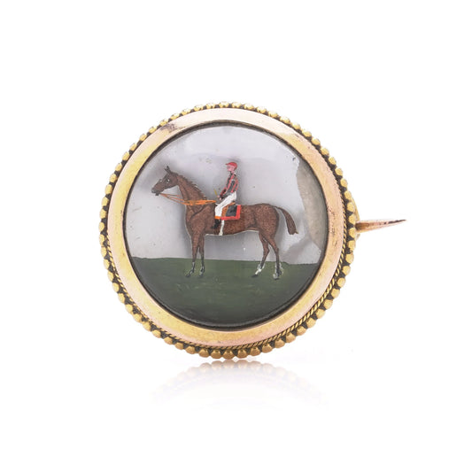A round crystal brooch in yellow gold featuring a jockey sitting on a horse's back, on a white background